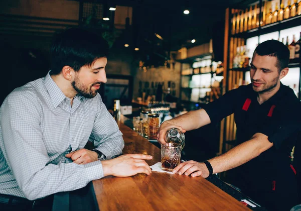 Müşteri barmans standında oturmuş ve nasıl barmen dökme bazı alkol cam için arıyorum. Bu sürecin kontrol ediyor. — Stok fotoğraf