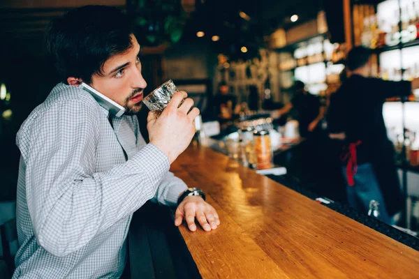 Young and dark-haired guy sitting at the waiters stand in the club and drinking some alcohol. Also he is talking on the phone and trying to drink at the same time. Barman stands far from him.