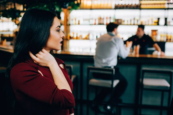 Krásný obrázek atraktivní bruneta sedí nedaleko stánku barmans. Dívá se na stranu kde je barman a jeho zákazníkem. Vypadá nadšený. — Stock fotografie