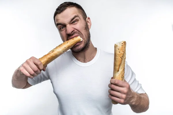 Zeer hongerig mens is honger. Hij brak een lang stuk stokbrood in twee stukken. Hij eet een van deze stukken. Hij kijkt brutale en raar. Geïsoleerd op witte achtergrond. — Stockfoto