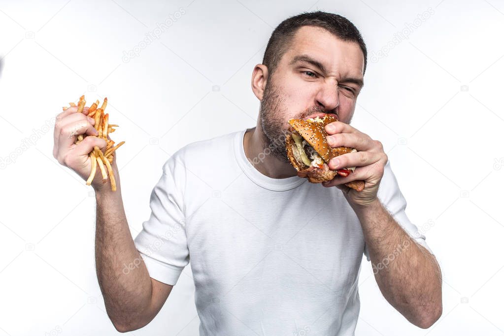 This guy is very delight of junk food. He is biting a big piece of burger and holding a full hand of french fries covered with ketchup. Young amn likes to eat oily meal. Isolated on white background.