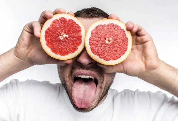 Смешная и хорошая фотография взрослого мужчины, который кладет две половинки грейпфрута себе на глаза. Также он показывает свой язык в камеру. Изолированный на белом фоне . — стоковое фото