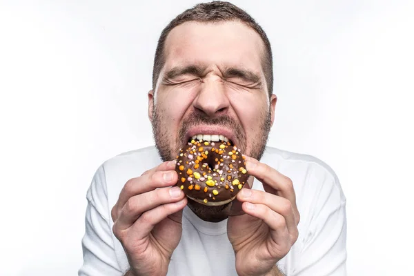 Este tipo se muere de hambre, así que está comiendo una gran dona de chocolate. Ha cerrado los ojos y disfrutando del momento. Al hombre Ypoung le gusta comer diferentes tipos de dulces. Aislado sobre fondo blanco . — Foto de Stock