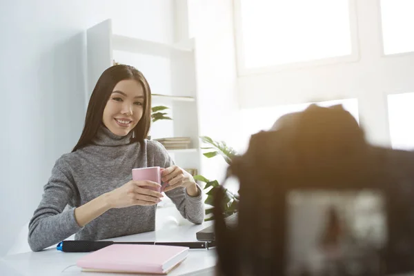 Nice and attractive girl is sittingat the table and drinking tea from pink cup. She is doing that in front of camera that recording it. She is looking to it and smiling. — Stock Photo, Image