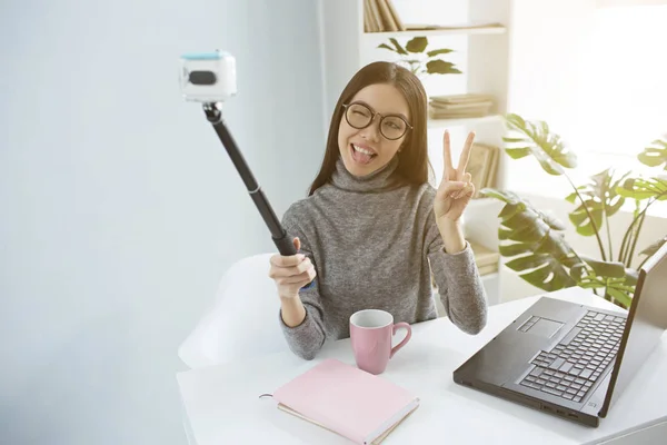 Die coole Brünette sitzt in einem hellen Raum und macht ein Selfie mit dem Selfie-Stick. Sie zeigt ein Stücksymbol vor der Kamera und schließt eines ihrer Augen. Mädchen sieht schön und liebenswert aus. — Stockfoto