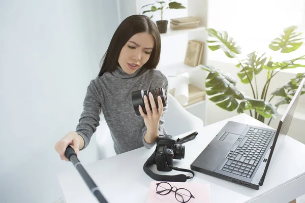 Professional blogger is sitting at the table and recording a video where she is looking to the cameras lens and studying it. She looks serious and cocncentrated. — Stock Photo, Image