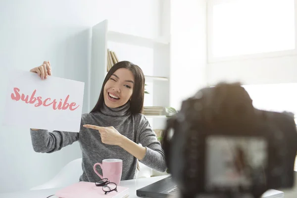 Uma fotografia de uma rapariga a gravar um vídeo na câmara. Ela tem um pedaço de papel com palavras impressas. Ela alsp aponta para o papel, o que significa que ela recomenda que todos se inscrevam em seu canal . — Fotografia de Stock