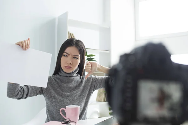 Angry female blogger is sitting at the table and recording video. She is looking to the paper she is holding. Girl is not pleased and happy. Also she is showing her big thumb down.