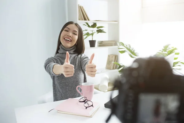 A picture of happy and delightul girl sitting at the table and recording her new vlog. She is showing her big thumbs up on her both hands and smiling. Young woman looks very good and nice. — Stock Photo, Image