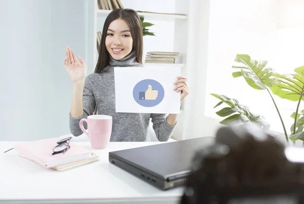 A picture of beautiful girl that likes when her followers like her video. She is sitting at the table and holding a picture with the like on it. She looks happy and pretty. — Stock Photo, Image