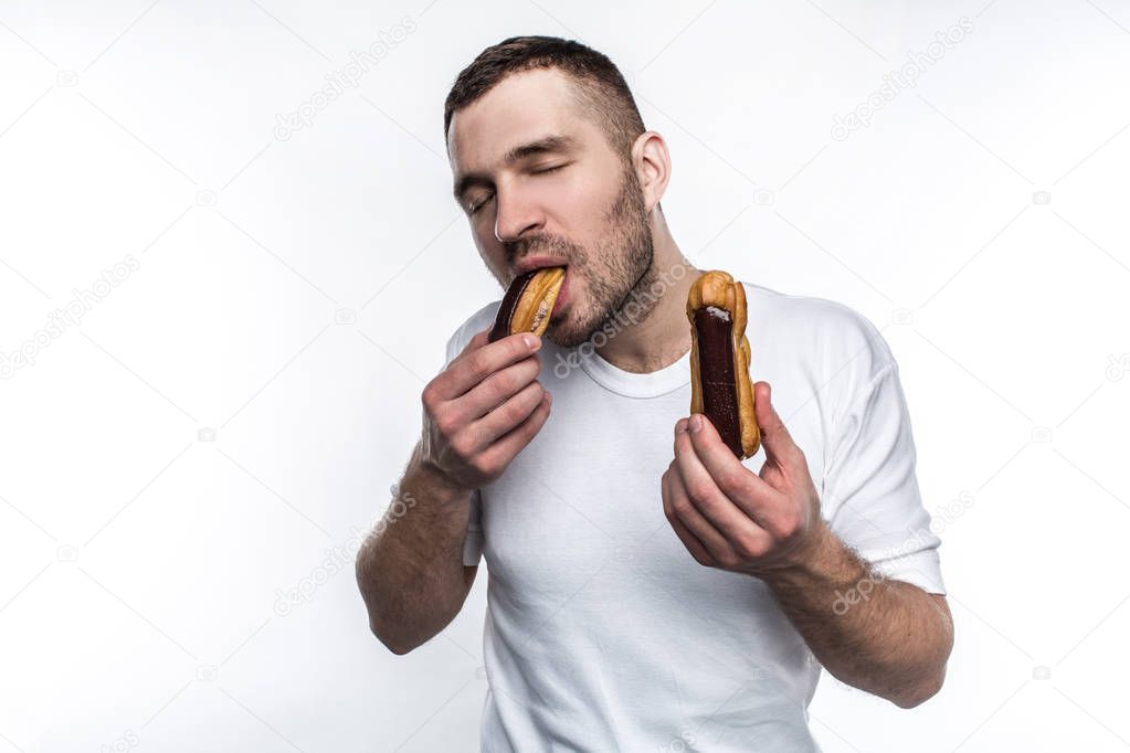 Strong and well-built guy is standing and eating eclair. Also he has another eclair in the other hand. Young man likes to eat oily fast food and sweet food as well. Isolated on white background.