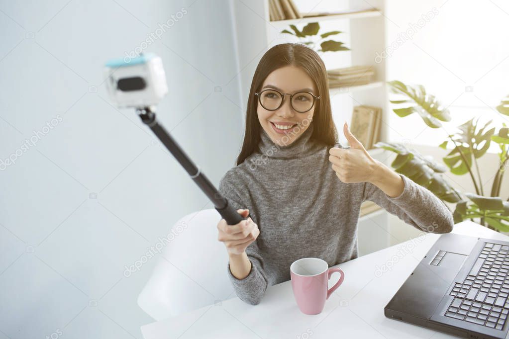 Beautiful blogger is sitting at the table in bright room and taking selfie using a selfie-stick for that. She is looking to camera and smiling.