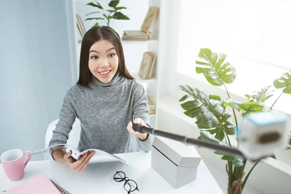 Selfie eines Mädchens, das am Tisch sitzt und ein Journal in der Hand hält. Mädchen blickt in die Kamera und lächelt. — Stockfoto