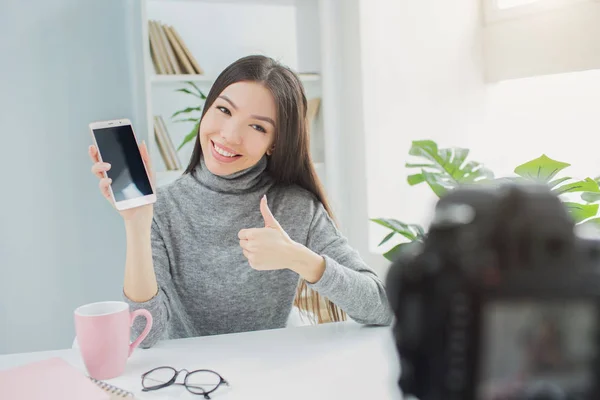 Fröhliche und coole Bloggerin zeigt das Telefon in die Kamera. Sie mag ihr neues Geschenk. junge Frau zeigt den großen Daumen nach oben. kann sie dieses Telefon ihren Abonnenten empfehlen. — Stockfoto