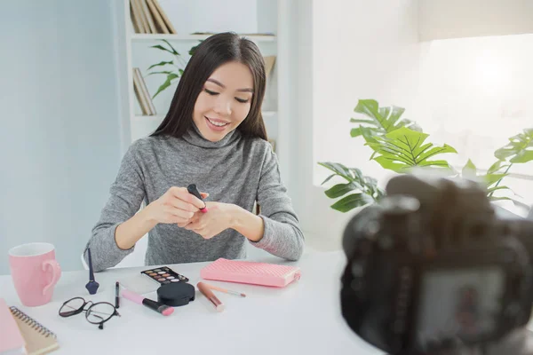 Beutiful girl is sitting at the table and making a video about her beauty products of the month. She is holding a lipstick and putting some of it on her hands to show the tone of it. — Stock Photo, Image
