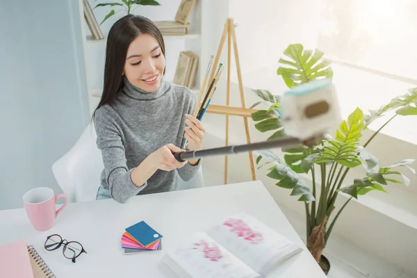 Blogger taking selfie with brushes. She is looking down to them and smiling. Girl is sitting at the table in a bright and large room. — Stock Photo, Image