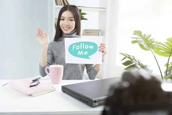 Nice brunette is showing a piece of paper where it is written follow me. Also she is waving to the camera and smiling. Girl is recording a video for heer channel. — Stock Photo, Image