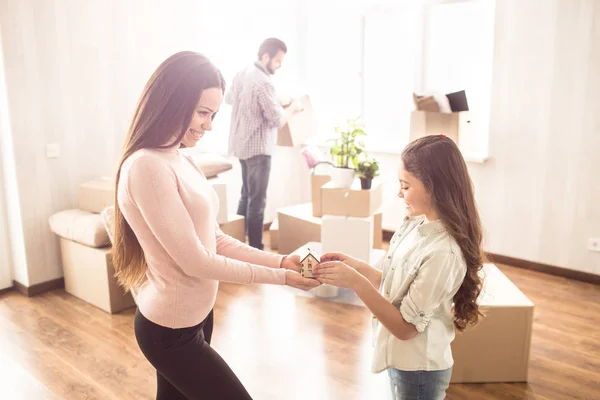 Glada mor och dotter står i ljusa rummet och hålla ett litet hus leksak som är gjord av trä. De ser att det och ler medan ungarna mannen uppackning grejer. — Stockfoto