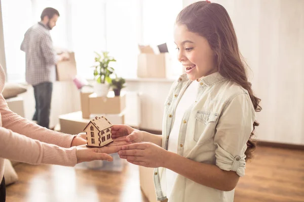 Vacker och glad liten tjej håller ett trä hus i händerna. Hon delar det med sin mor. Fadern håller en låda med grejer som behöver packas upp. — Stockfoto