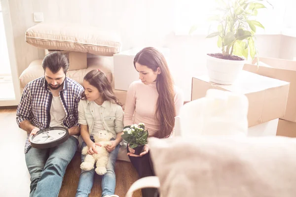 Nice picture of parents with their child sitting on floor in room and looking to clock that man holds. He wants to make it work. Young woman holds a jar with plant in her hands.