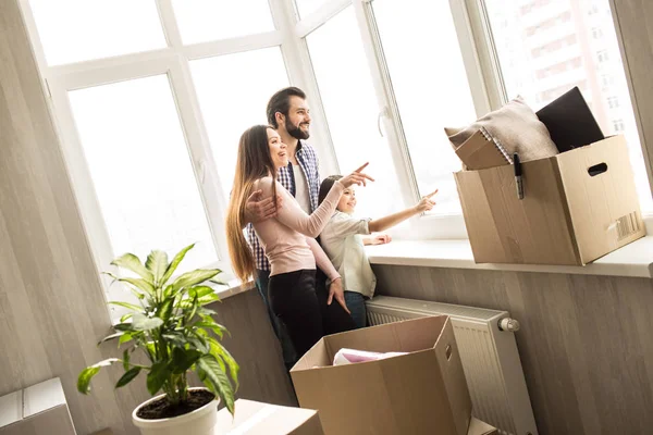 Man, kvinna och barn står framför det stora fönstret och titta utanför. Kvinna och barn pekar på fönstret. De ser glada. Förutom dem står man. Det finns lådor med grejer. — Stockfoto