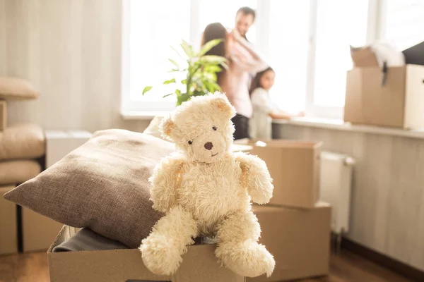 Immagine carina di giocattolo bianco di orso seduto sulla scatola con cuscini. Sta guardando dritto davanti a noi. C'è anche una famiglia nella stanza. Guardano fuori dalla finestra e parlano . — Foto Stock