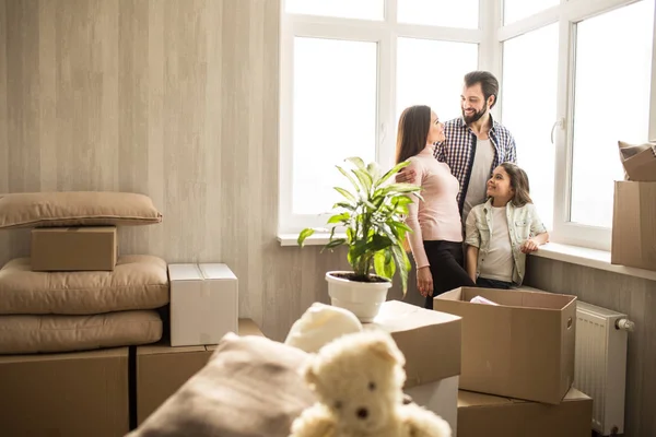 Bonito y luminoso apartamento. La familia joven y feliz acaba de mudarse a ella. Hay cajas con sus cosas por todas partes. La familia está de pie cerca de las ventanas y hablando entre sí. . — Foto de Stock