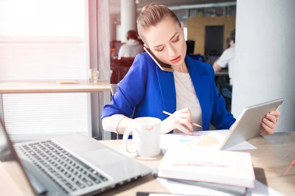 Girl in blue jacket is sitting at the table and working. She is talking on the phone and writing down information. Also she has a tablet in her hands. This woman is busy. — Stock Photo, Image