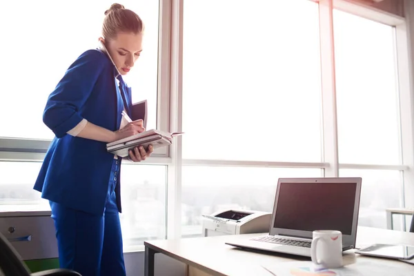 Vacker kvinna i blå kostym är stående i sitt kontor och prata i telefon. Hon skriver ned information. Det finns även laptop och cup på hennes bord. — Stockfoto