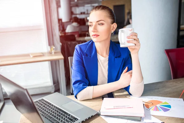 Nice and attractive girl is sitting on the table and drinking tea. She is looking to window and dreaming. This adult has some rest after working with papers and documents.