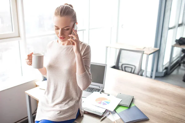 Business woman is standing at her office table and talking on the phone. She is holding a cup of tea in one hand and holding the phone with the other one. There are lots of notebooks on the table. — Stock Photo, Image