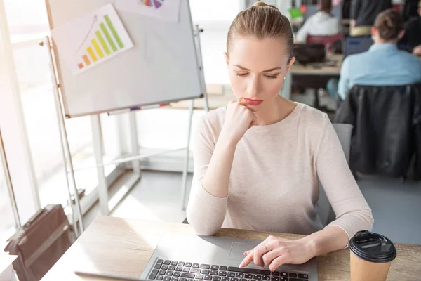 Detta professional är sitter vid bordet och ser att skärmen bärbara datorer. Hon studerar. Flicka har en kopp kaffe på bordet. Hon ser allvarlig och eftertänksam. — Stockfoto