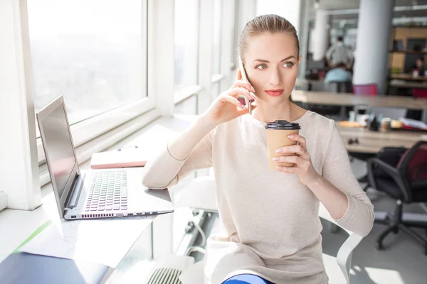 Девушка разговаривает по телефону. Она смотрит направо и держит чашку кофе близко ко рту. Женщина отдыхает после продуктивной работы . — стоковое фото