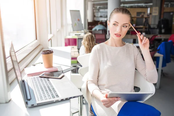 Allvarlig och eftertänksam ung kvinna tänker. Hon tittar till höger. Hon har också en tablett i händerna och en bärbar dator på sitt bord. — Stockfoto