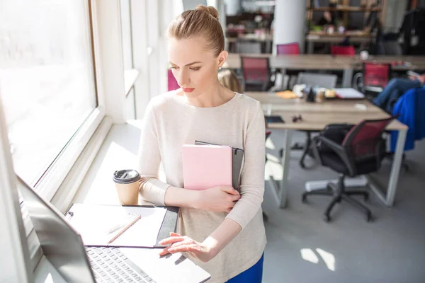 Allvarliga och upptagen kvinna är ser att den bärbara datorn och använder den. Hon innehar också några anteckningsböcker i händerna. Kvinnan är arbeta. — Stockfoto