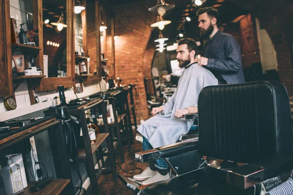 Zdjęcie z daleka. Facet jest siedzi na krześle i przygotowuje się do cięcia brodę. Jego fryzjerka jest gotowy do rozpoczęcia pracy. — Zdjęcie stockowe