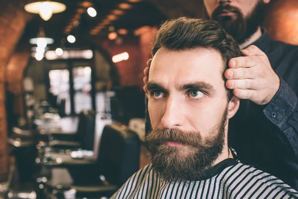 Close-up de cara sério e barbudo sentado na cadeira e olhando para a frente. Seu cabeleireiro está modelando seu cabelo . — Fotografia de Stock