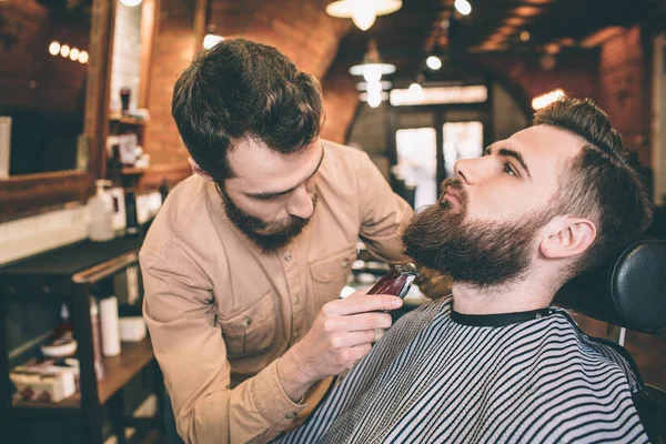 Dois tipos sérios estão numa barbearia. Um deles é o cliente e sentado em uma cadeira, enquanto o cabeleireiro está fazendo alguma magia, cortando um pouco da barba . — Fotografia de Stock