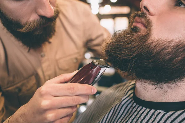 Feche e corte a vista de um cara que está cortando alguma barba de seu cliente com um raser elétrico. Ele está fazendo isso muito preciso e gentil . — Fotografia de Stock