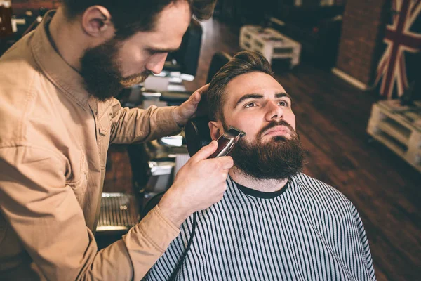 Cabeleireiro agradável e bom está cortando alguns dos clientes barba com um razer elétrico. Ele está a fazer isso com muita precisão. . — Fotografia de Stock