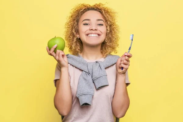 Glückliches und zufriedenes Mädchen lächelt. In der einen Hand hält sie einen grünen Apfel, in der anderen eine Zahnbürste. Sie sorgt sich um ihre Zahngesundheit. isoliert auf gelbem Hintergrund. — Stockfoto