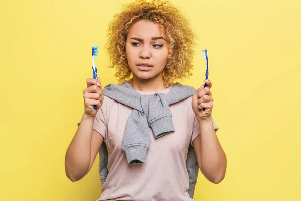 Fille tient deux brosses à dents dans ses mains et regarde l'un d'eux. Sa vue est suspecte. Elle ne sait pas quelle brosse à dents utiliser. Isolé sur fond jaune — Photo