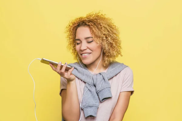 Happy girl is holding a phone in her hand. She is listening to music. Young woman is enjoying the moment. Isolated on yellow background.