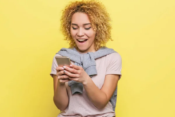 Glückliches Mädchen blickt auf den Bildschirm des Telefons und lächelt. Sie ist aufgeregt und glücklich. isoliert auf gelbem Hintergrund. — Stockfoto