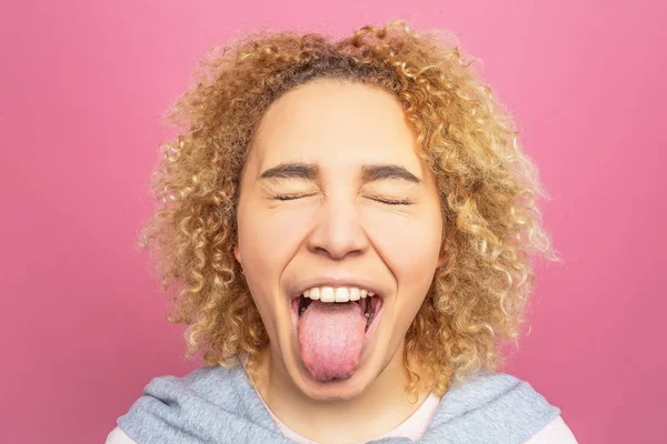 Close up of girls face. She is showing her big tongue and keeping her eyes closed. This girl behaves like a child. Isolated on pink background.