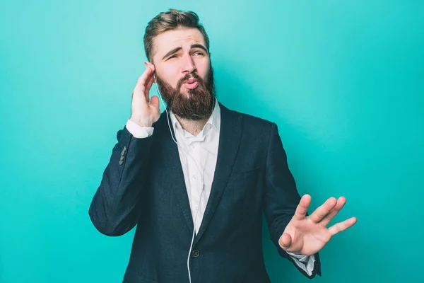 Guy in suite is staanding and listening to the music on the phone. He is enjoying the moment and looking to the right. He is holding right hand close to the ear. Isolated on blue background.