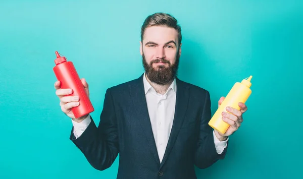 Atraktivní muž v sadě drží dvě lahve ve svých rukou. Jedním z nich je s kečupem a dalším z nich je s hořčicí. Tyto láhve jsou barevné. Izolované na modrém pozadí. — Stock fotografie