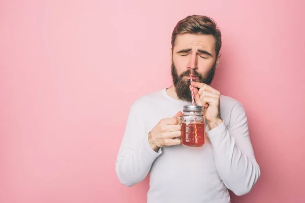 Skäggig kille i en vit tröja är att dricka lite te från cup men halmen. Han njuter nu. Isolerad på rosa bakgrund. — Stockfoto