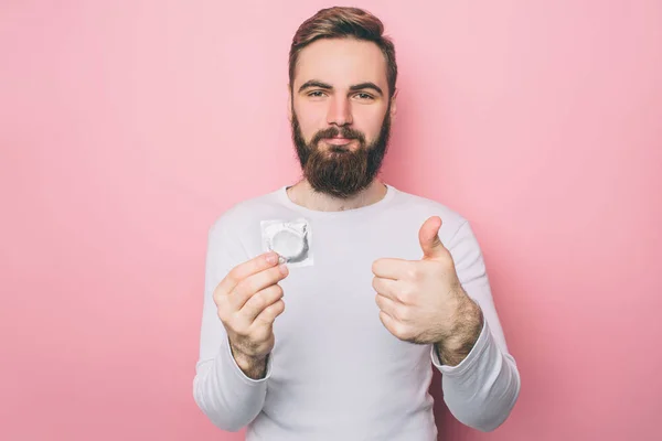 Stilig man håller en kondom i handen. Även visar han sin stora tummen. Han föredrar att skydda sin hälsa. Isolerad på rosa bakgrund. — Stockfoto