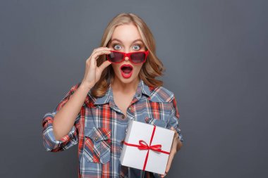 Sürpriz kız bir hediye onun elinde tutuyor. O diddnt bekliyoruz. Genç Bayan düz ön seyir ve gözlüklerini burnuna tutarak. Gri arka plan üzerinde izole.